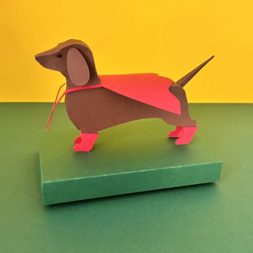 Kit de paper art pour créer un teckel et ses accessoires. Ici, il a revêtu sa cape et ses bottes de super-héros