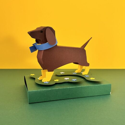 Kit de paper art pour créer un teckel et ses accessoires. Ici, il a revêtu des bottes jaunes et une écharpe bleue